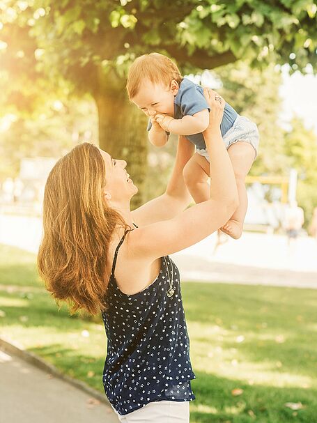 Mother lifting her smiling baby in the air outdoors, both enjoying a sunny day with green trees in the background.