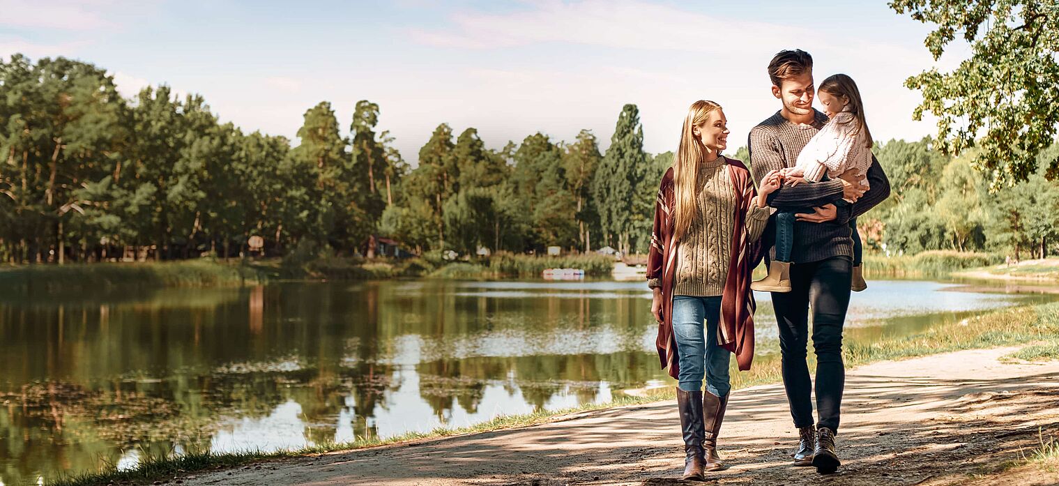 Family of four walking along a path by a lake, with trees and clear sky in the background, enjoying a peaceful outdoor moment.