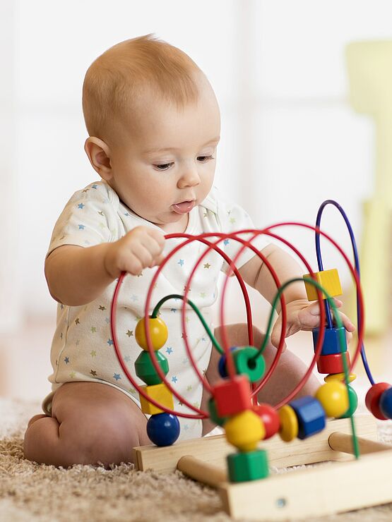 Baby playing with a colorful bead maze toy while sitting on a carpeted floor in a brightly lit room.