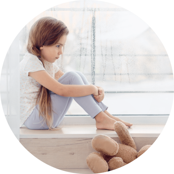 A young girl sitting on a windowsill with a teddy bear on the floor, looking thoughtful and slightly sad