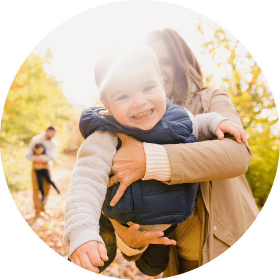 Happy baby in a blue outfit being held by a smiling adult in a sunlit, autumn park, with another person walking in the background