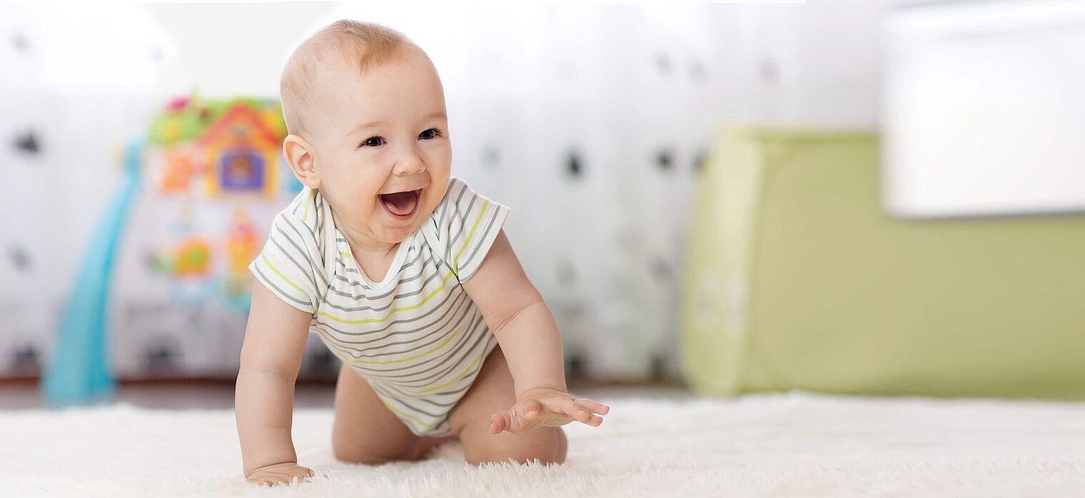 "Happy baby in a striped onesie crawling on a soft carpet in a brightly lit room, with toys and furniture in the background.