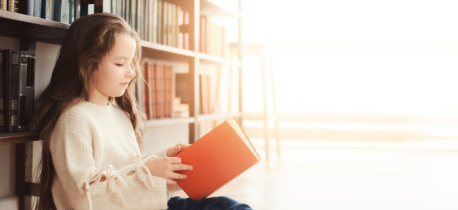 A young girl in a library reading a book with a calm and focused expression.