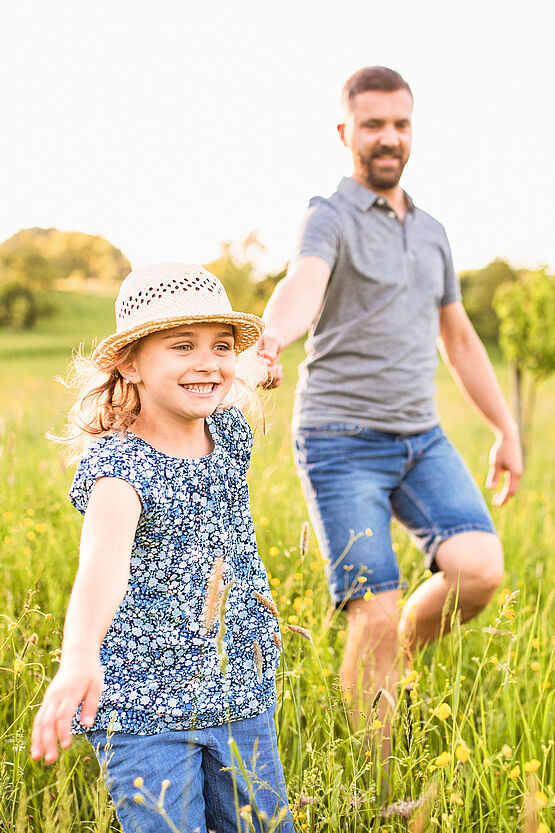 Smiling young girl wearing a straw hat and a blue floral dress, running through a sunny field with a man, likely her father, following behind