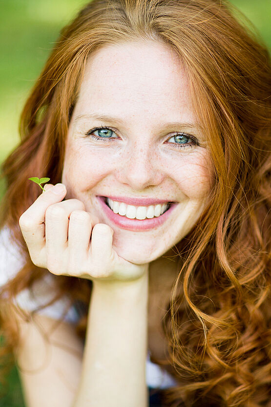 "Close-up of a smiling red-haired woman lying on grass, with her head resting on her hand."