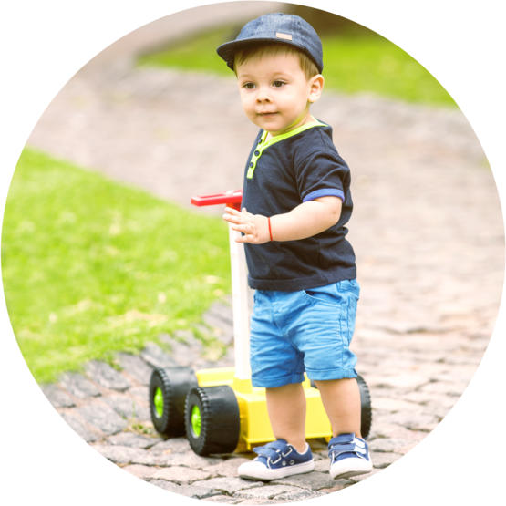 Young toddler wearing a cap and casual clothes, pushing a small yellow push toy on a paved path in a park.