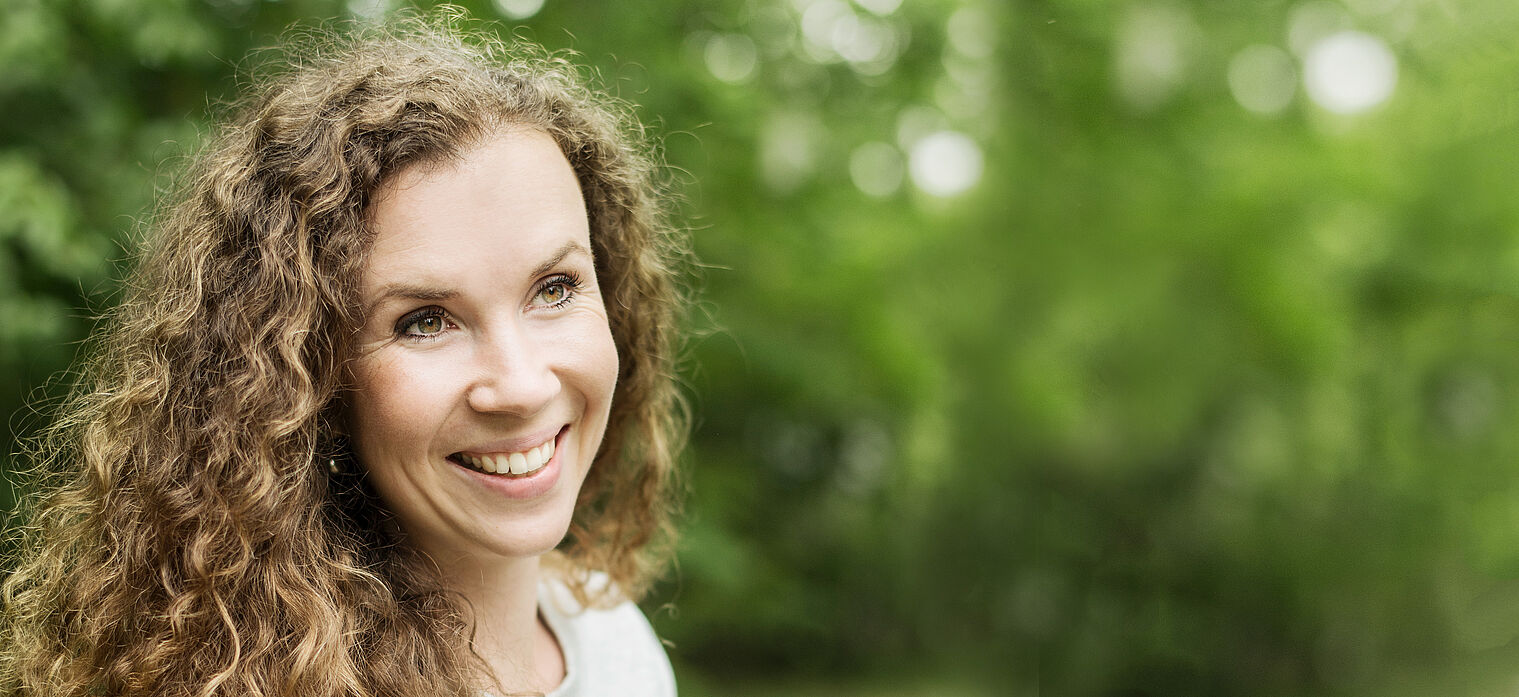 Smiling woman with curly hair standing outdoors in a green, leafy environment.