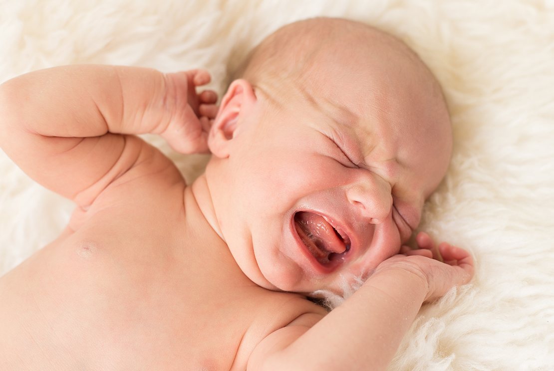 A newborn baby lying on a soft white blanket, crying with eyes closed and hands near the face.